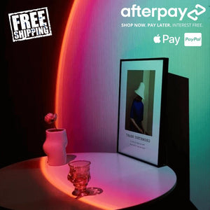 Sunset lamp casting a vibrant gradient of pink, orange, and green light on a wall, illuminating a modern vanity or desk setup with ambient glow.