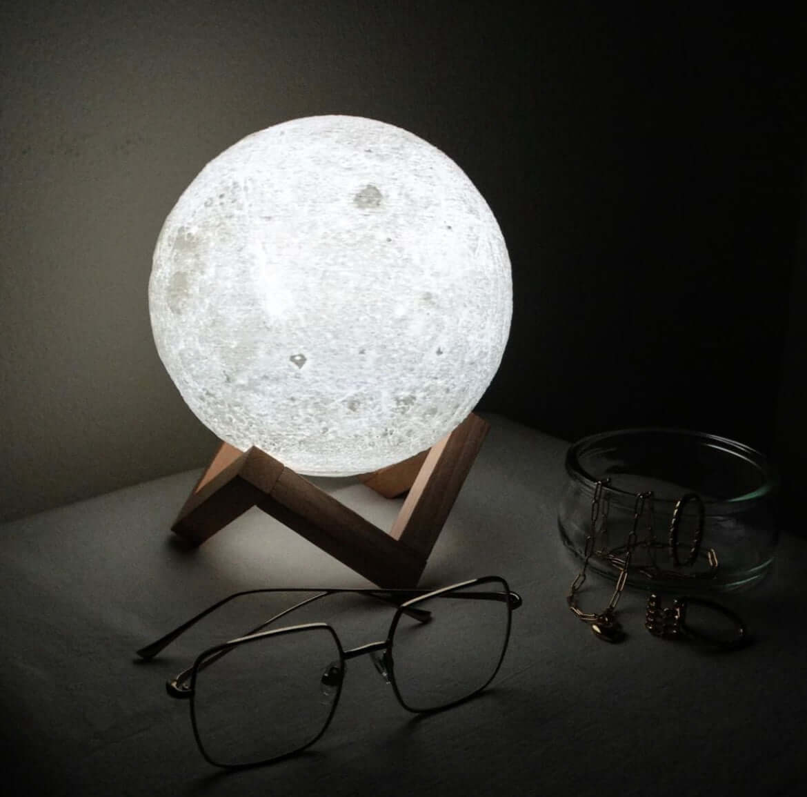 Bright white moon lamp with realistic lunar surface detail, glowing on a wooden stand in a dark room with string lights nearby.