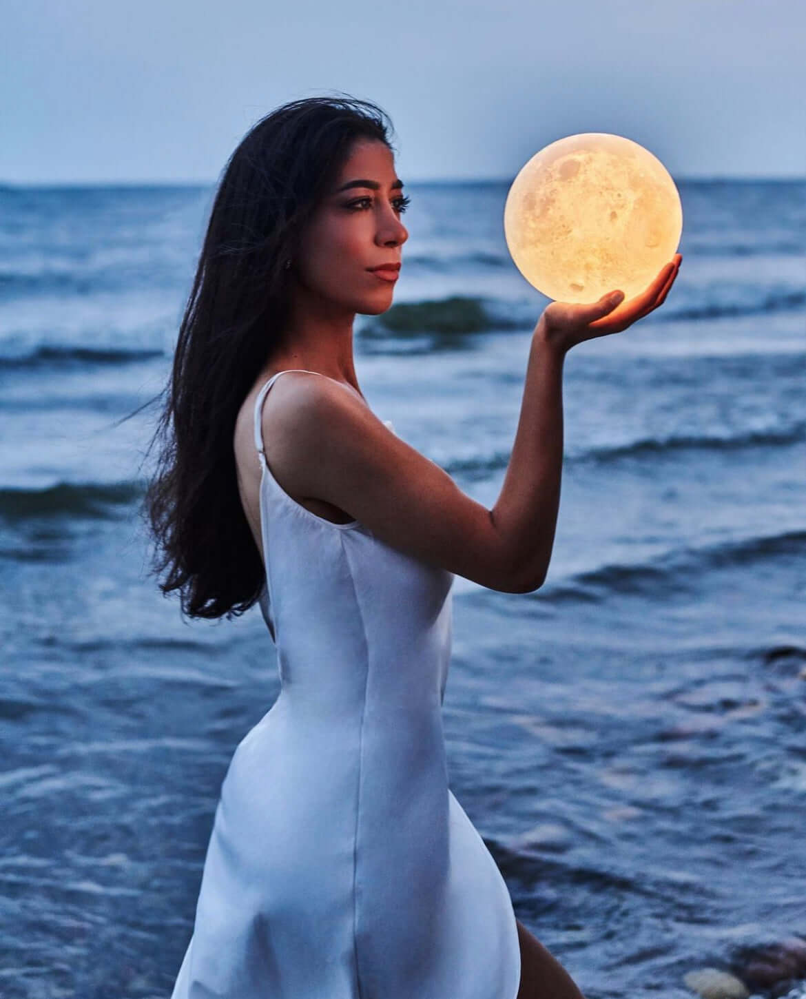 Woman in a white dress standing by the ocean at sunset, holding a glowing moon lamp that appears like a real moon in her hand.