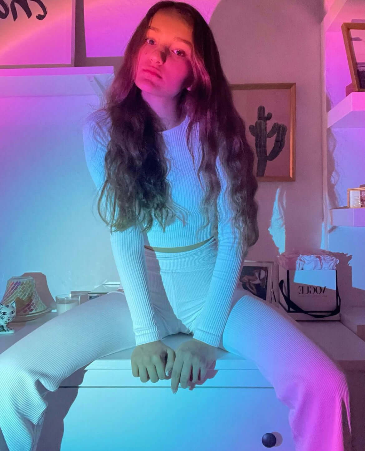 Young woman sitting on a bed in a room lit by a sunset lamp, surrounded by pink, blue, and purple gradient lighting for a dreamy, aesthetic vibe.