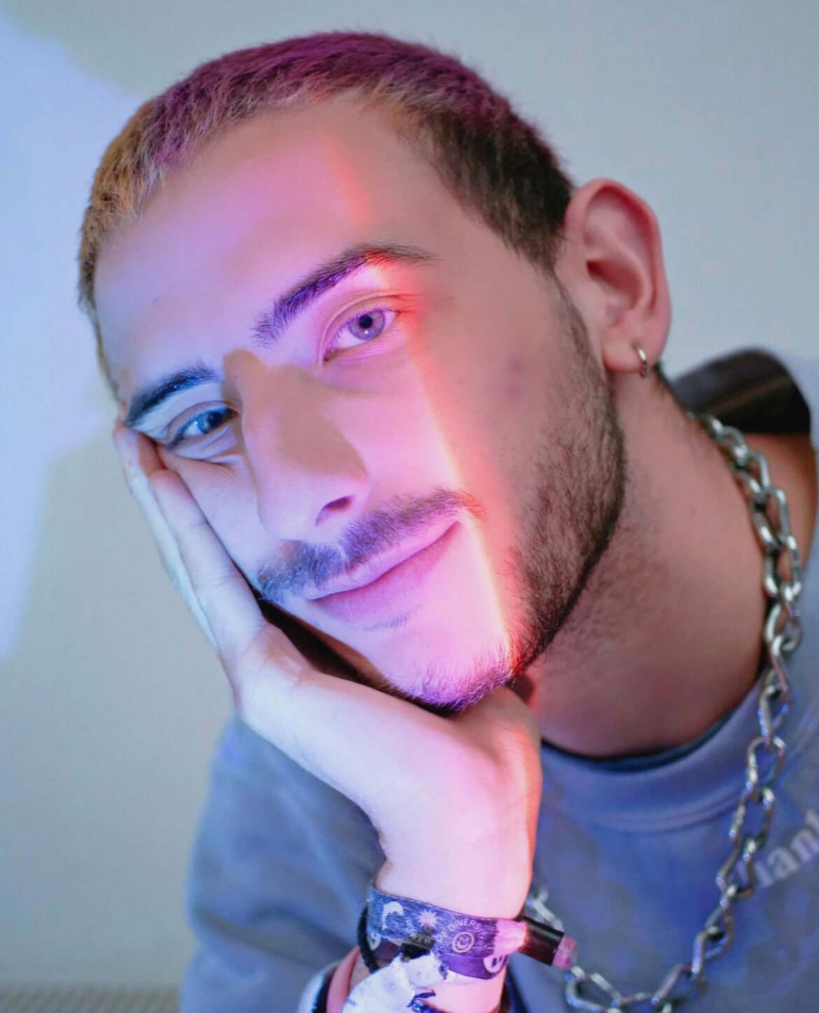 Smiling man resting his head on his hand with soft purple and blue lighting from a sunset lamp casting a colorful glow on his face.