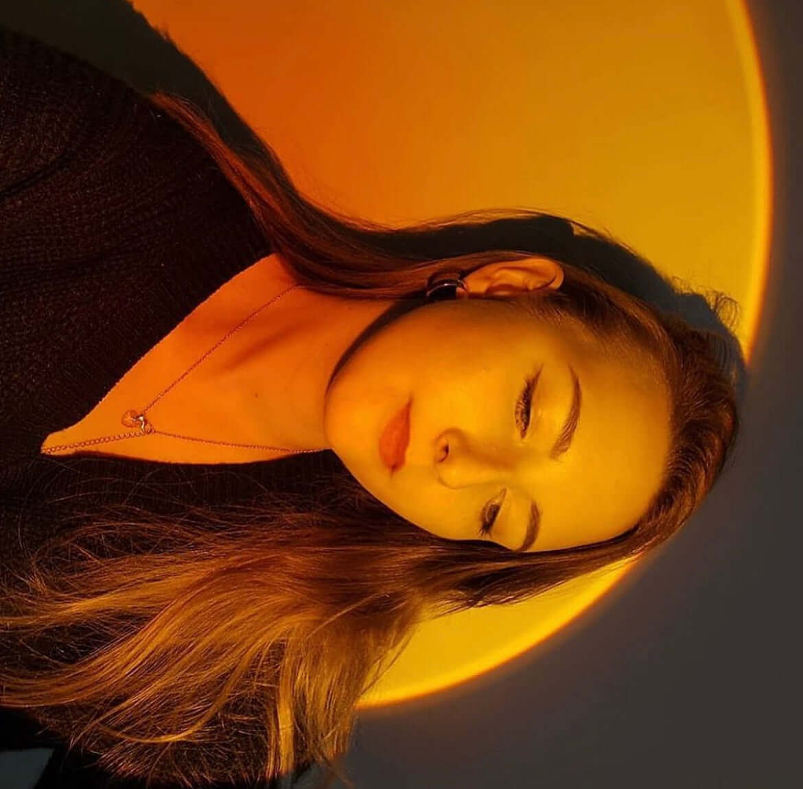 Woman peacefully resting in front of a wall illuminated by a sunset lamp, with a warm orange glow creating a calm and dreamy ambiance.
